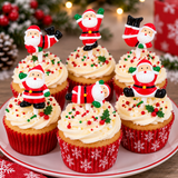 Six decorated cupcakes on a white plate with red snowflake trim, topped with white frosting and colorful holiday sprinkles, each featuring a Santa Claus figure on a stick. The cupcakes are set against a softly blurred festive background with Christmas trees and warm lights.