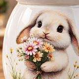 Close-up of a glossy white ceramic jar featuring a detailed illustration of a fluffy cream-colored bunny holding a bouquet of pink, yellow, and white flowers.