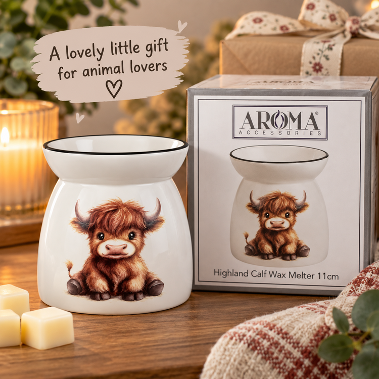 A white ceramic highland calf wax warmer with a printed illustration of a fluffy brown calf, shown next to its retail box. The scene is styled on a wooden table with a lit candle, cream wax cubes, and a cozy knitted blanket in the foreground, with a text overlay reading 'A lovely little gift for animal lovers'.