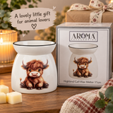 A white ceramic highland calf wax warmer with a printed illustration of a fluffy brown calf, shown next to its retail box. The scene is styled on a wooden table with a lit candle, cream wax cubes, and a cozy knitted blanket in the foreground, with a text overlay reading 'A lovely little gift for animal lovers'.