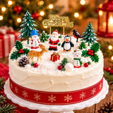 A decorated Christmas cake on a white pedestal stand, topped with festive figurines including Santa Claus, a penguin, snowmen, a robin, a deer, pinecones, holly, miniature trees, and a gold 'Merry Christmas' sign. The cake has white frosting, a red ribbon with gold snowflakes, and is set against a warm, blurred Christmas tree and lantern background.