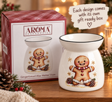 A white ceramic gingerbread wax warmer with a smiling gingerbread man illustration, shown next to its retail box. The scene is styled on a wooden surface with holiday greenery, pinecones, and warm blurred lights in the background.