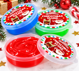 Three round plastic tubs of Christmas-themed slime arranged on a white marble surface with holiday decorations. One tub is blue and one is green, and one clear tub in front contains bright red slime.