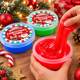 Three round plastic tubs of Christmas slime on a wooden table, with a hand stretching the red slime in the front tub. The lids show colorful holiday labels reading 'Happy Christmas Christmas Slime.