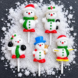 Five festive snowman topper decorated in holiday colours, arranged on a bed of white faux snow against a dark textured background.