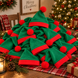 A pile of plush Christmas elf hats in green and red, styled on a holiday table with a lit Christmas tree, pinecones, a candle, and a 'Merry Christmas' sign in the background.