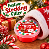 A round red plastic tub of Christmas-themed slime sits on a wooden surface surrounded by festive holiday props, including a Santa hat, candy canes, wrapped gift boxes, pine branches, and a large decorative sign reading 'Festive Stocking Filler.' The lid label says 'Merry Christmas Christmas Slime' with Santa and a reindeer illustration.