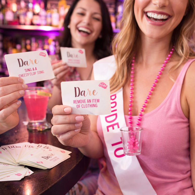 Two women at a bar with 'Dare' cards and a sash, likely participating in a bachelorette party game.