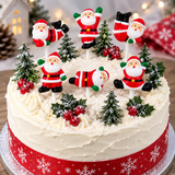A round white-frosted cake decorated with six Santa Claus figurines, small evergreen trees, holly leaves, and red berries, with a red ribbon around the base.