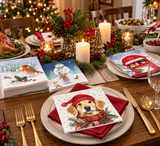 A beautifully decorated Christmas table setting featuring stacks of printed paper napkins with festive holiday illustrations, including a robin, a snowman, and a golden retriever in a red Santa hat. The scene is styled with gold flatware, candles, pinecones, and holiday greenery on a wooden dining table.
