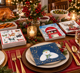 A festive holiday table setting featuring stacks of printed paper napkins with Christmas illustrations, including a snowman on blue with gold stars, a red post office scene, and a reindeer on white. The napkins are styled on plates with gold cutlery on a decorated dining table with candles, a roasted turkey, and holiday greenery.