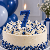 A frosted birthday cake with white piped frosting, blue and white sprinkles, and a glittery blue number 7 candle on top, photographed in a decorated party setting with blue balloons in the background.