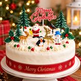 A frosted Christmas cake on a white cake stand, decorated with Santa Claus, a snowman, a penguin, a reindeer, a robin, pine trees, holly, and a red 'Merry Christmas' topper. The cake has a red ribbon with gold snowflakes and 'Merry Christmas' text, set against a blurred festive background.