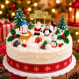 A decorated Christmas cake on a white pedestal stand, topped with festive figurines including Santa Claus, a penguin, snowmen, a robin, a deer, pinecones, holly, miniature trees, and a gold 'Merry Christmas' sign. The cake has white frosting, a red ribbon with gold snowflakes, and is set against a warm, blurred Christmas tree and lantern background.
