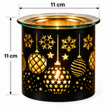 A black cylindrical wax warmer with a festive holiday cutout pattern featuring gold snowflakes, stars, and hanging ornaments. The top rim is clear glass, and a warm light glows through the cutouts.