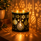 A black holiday-themed candle holder with laser-cut snowflakes, stars, and ornaments, glowing warmly on a wooden table and casting decorative shadows on the wall.