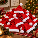 A large pile of red velvet Santa hats with white trim and white pom-poms arranged on a wooden table, with a Christmas tree, ornaments, greenery, and a lit candle in the background.