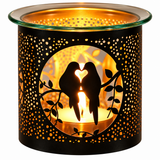 A black metal candle warmer with a glass rim, featuring a cutout design of two birds on a branch with a heart-shaped flame in the center, glowing warmly against a white background.
