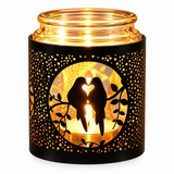 A black metal candle holder with a warm glowing LED light inside, featuring a silhouette cutout of two birds forming a heart shape on a branch, surrounded by a dotted pattern.