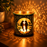 A black metal candle holder with a cutout silhouette of two birds forming a heart shape, lit with a warm candle inside and casting a tree-like shadow on a wooden surface and wall.