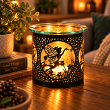 A black metal tea light candle holder with a fairy silhouette and floral cutout pattern, glowing warmly on a wooden table in a cozy living room setting.