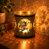 A black metal fairy-shaped candle holder with floral cutouts, glowing with warm yellow light on a wooden table, casting a fairy shadow on the wall. A potted plant sits in the background.
