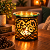 A black metal scented candle holder with a heart-shaped cutout and scrollwork pattern, glowing with warm light on a wooden table in a cozy living room setting.