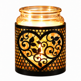 A lit glass jar candle holder with a black metal heart-shaped cage featuring scrollwork and leaf cutouts, casting a warm golden glow against a white background.