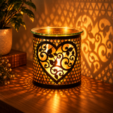 A black metal heart-shaped candle holder with intricate scrollwork cutouts, illuminated from within by a warm candlelight. The holder is placed on a wooden table, casting decorative shadows on the wall and surface, with a potted plant in the background.