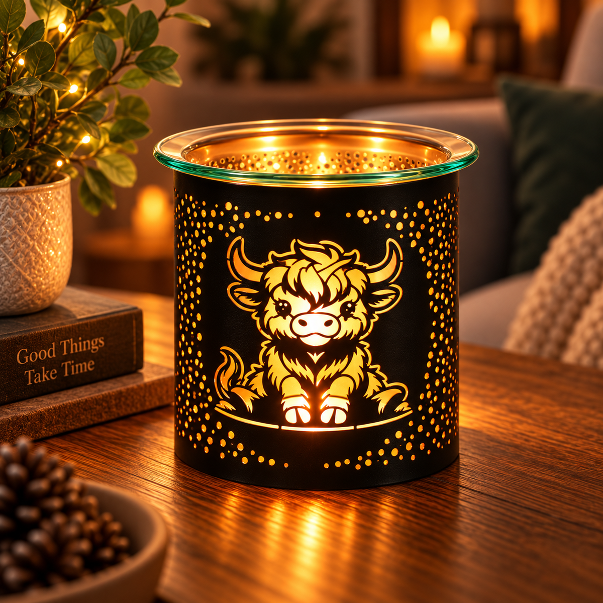 A black cylindrical candle holder or diffuser with a glowing warm light, featuring a cutout illustration of a fluffy yellow-and-white cow and gold speckled dots. It sits on a wooden table in a cozy indoor setting with warm ambient lighting, a plant, and books in the background.
