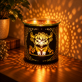 A black metal scented candle holder with a glowing yellow bull design, placed on a wooden table casting warm golden light patterns on the wall and surface. A potted plant is visible in the background.