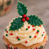 A close-up of a frosted cupcake topped with white icing, red and green sprinkles, and a green holly leaf decoration with red berries, set against a softly blurred neutral background.