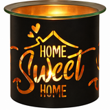 A black metal candle warmer with a glass top rim, illuminated from within to show a warm orange glow. The front features a cutout design with the words 'HOME Sweet HOME' and a house outline with hearts.