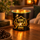 A black metal candle holder with a clear glass rim sits on a wooden table, glowing warmly from within. The front is printed with the phrase 'HOME Sweet HOME' in gold lettering.