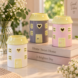 Three small house-shaped ceramic holders in white, lavender, and pink, each with pale yellow roofs and door details, arranged on stacked books with flowers in the background.