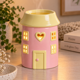A pink and pale yellow house-shaped ceramic oil burner with glowing windows and a heart-shaped cutout on the front, set on a tabletop with a plant and books in the background.