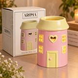A pink house-shaped wax warmer with a pale yellow roof and door, shown next to its branded retail box on a wooden surface. The warmer has cutout windows and a heart-shaped window, with warm light glowing from within.