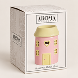 A pink house-shaped wax warmer with a pale yellow roof and door, shown in front of its branded retail box. The box reads AROMA ACCESSORIES and House Wax Melt 12cm.