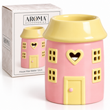 A pink and yellow house-shaped wax warmer with a heart-shaped window and cutout details, shown next to its white AROMA ACCESSORIES retail box.