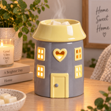A ceramic house-shaped wax warmer with a gray body and pale yellow roof, door, and window frames, shown on a wooden table with candles, books, and plants in a cozy indoor setting.