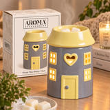 A house-shaped wax warmer by Aroma Accessories in gray and yellow, shown with its retail box on a wooden table. The warmer has a yellow roof, yellow door, and yellow window cutouts, with visible steam rising from the top.
