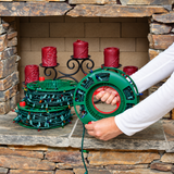 A person's hands are shown holding a green spool of Christmas string lights in front of a stone fireplace. More spools of green and red string lights are stacked on the hearth next to a black wrought-iron candle holder with red pillar candles.