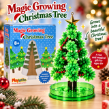 Magic Growing Christmas Tree toy set by PlayWrite, showing the green plastic artificial tree on a round green base next to its blue retail box, with festive holiday decorations and warm blurred lights in the background.