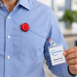 A close-up of a person wearing a light blue button-down shirt, with a red retractable badge reel clipped to the chest pocket. The person is holding a clear ID badge with a white nameplate that reads Aim2Entertain and Michael Carter, Operations Manager, against a softly blurred office background.
