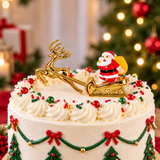A decorated white frosted cake topped with a gold reindeer and Santa Claus sleigh decoration, with red and green candy sprinkles and a blurred Christmas tree in the background.