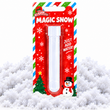 A plastic magic snow tube in its festive red holiday blister packaging, displayed on a bed of white faux snow. The packaging features 'Merry Christmas MAGIC SNOW' text, candy cane stripes, a snowman illustration, and instructions to 'JUST ADD WATER!'.