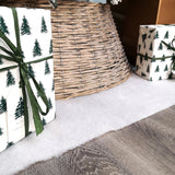 Two wrapped presents with green tree patterns on a white fake snow surface next to a woven basket.