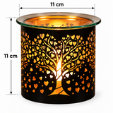 A black metal candle holder with a laser-cut tree design filled with small hearts, glowing with warm orange light from an internal candle. The top rim is clear glass, and the image includes dimension overlays showing a height and width of 11 cm on a white background.