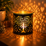 A black metal candle warmer with a tree of life cutout design and heart-shaped patterns, lit with a warm glow that casts heart-shaped shadows on a wooden table and wall.