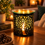 A black metal candle holder with a tree silhouette cutout, lit with a warm glowing candle inside, set on a wooden table in a cozy living room scene.
